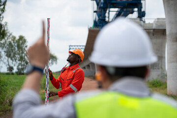 The survey team is operating a theodolite while referencing road construction plans. Civil engineers are taking measurements with surveying equipment at the construction site.