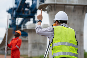 The survey team is operating a theodolite while referencing road construction plans. Civil engineers are taking measurements with surveying equipment at the construction site.