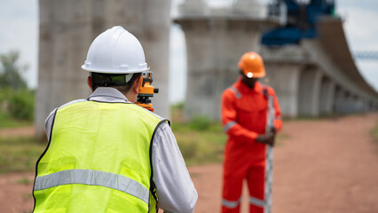 The survey team is operating a theodolite while referencing road construction plans. Civil engineers are taking measurements with surveying equipment at the construction site.