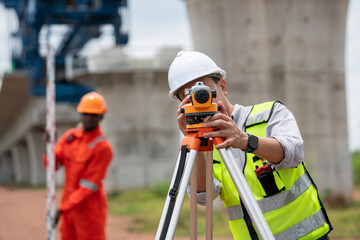 The survey team is operating a theodolite while referencing road construction plans. Civil engineers are taking measurements with surveying equipment at the construction site.