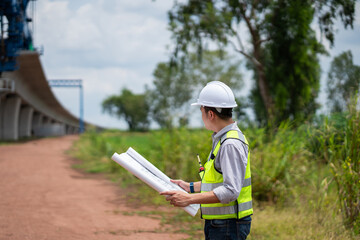 The survey team is operating a theodolite while referencing road construction plans. Civil engineers are taking measurements with surveying equipment at the construction site.