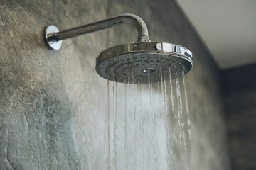 A modern, gleaming chrome showerhead releases a steady cascade of fresh, clear water, creating an inviting and refreshing scene. Set against a textured, contemporary grey wall, this image evokes a str