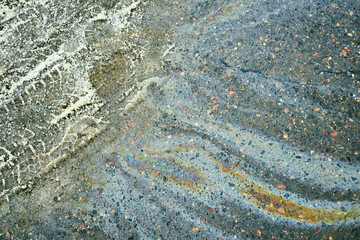Oil spill on asphalt path forming colorful rainbow pattern near snow and tire tracks, pollution concept