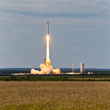 powerful space rocket launch with massive exhaust plumes rising from a launchpad into the blue sky over a green field and forest landscape.