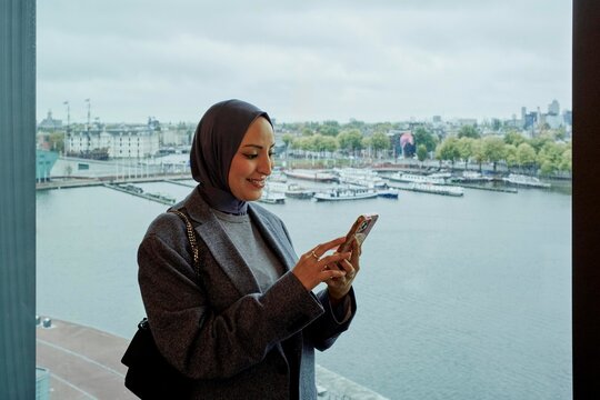 Smiling Woman in Hijab Texting by Waterfront in Urban Setting