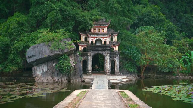 A steady, low-angle shot of the iconic triple-arched stone gate of Bich Dong Pagoda, framed by lush greenery and a calm lotus pond at the base of a limestone mountain.