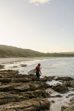 Young woman in a wetsuit walking over rocks to enter the ocean