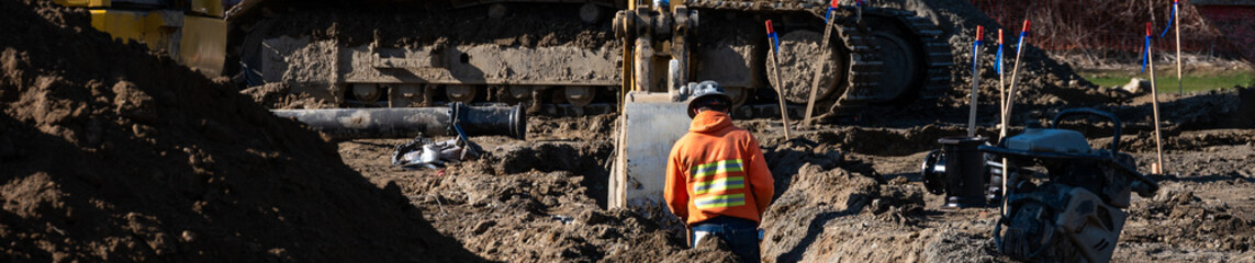 Construction worker standing in a dirt trench while excavator fills in dirt to cover a newly...