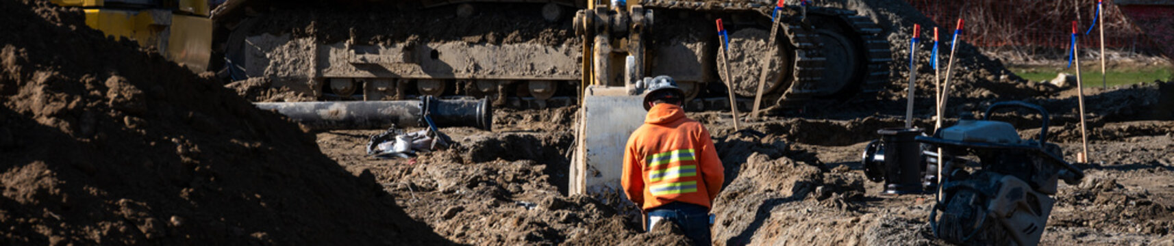 Construction worker standing in a dirt trench while excavator fills in dirt to cover a newly installed water pipeline, new municipal water distribution utility project work
