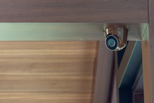 A security camera is mounted on a wooden shelf. The camera is black and has a lens. The shelf is made of wood and has a brown color.