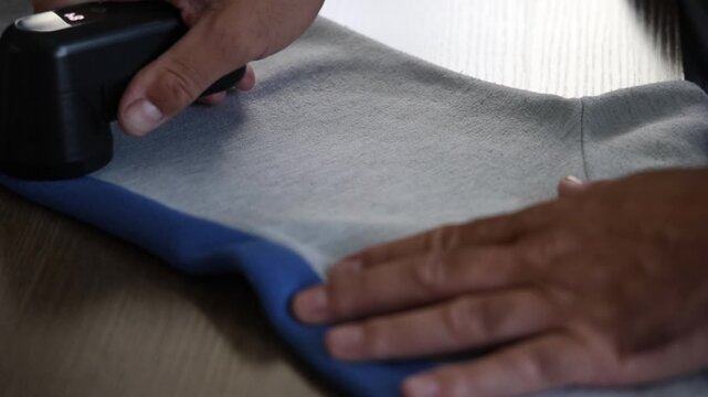Close up of person using electric fabric shaver to remove lint and pills from grey sweater. Handheld device cleaning clothing surface indoors in soft natural light with selective focus.