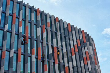 Obraz premium Window washers cleaning windows on modern office building facade. High rise window cleaners using rope access. Industrial alpinists washing exterior of skyscraper. Dangerous risky work at height