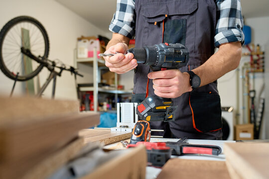 Carpenter in workwear holding cordless drill over workbench with wood boards and tools. Concept of woodworking DIY repair and furniture assembly