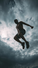 A man is jumping in the air with his arms outstretched. The sky is cloudy and the man is wearing shorts