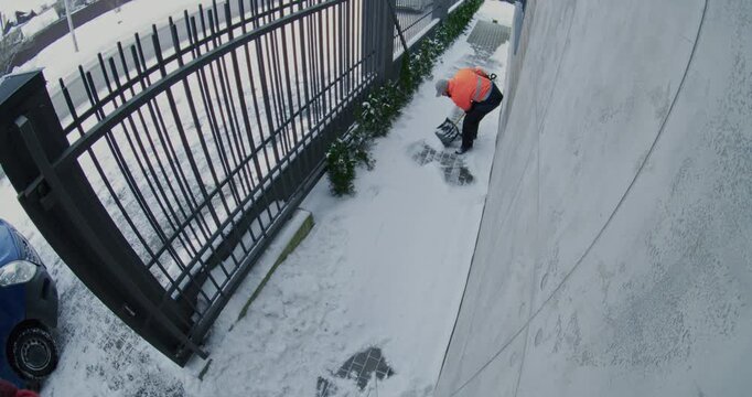 Male Utility Worker Wearing Reflective Jacket Removes Snow From Paved Walkway Beside Fence During Winter Conditions. Safe Pedestrian Access And Seasonal Maintenance Captured By Surveillance Camera.