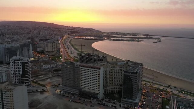 Aerial wide shot capturing the curve of Tangier Bay at sunset. Features modern high-rise buildings along the Corniche, the sandy beach, and the calm Mediterranean sea in Northern Morocco.
