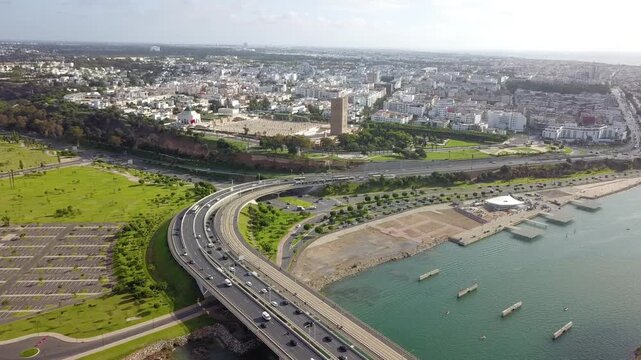 Aerial shot highlighting the sweeping curve of the Rabat-Sal&eacute; bridge. Captures busy vehicle traffic leading the eye towards the historic Hassan Tower and the city's lush green landscapes.