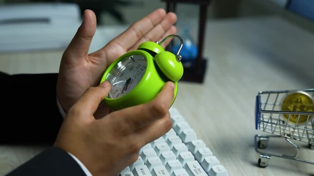 Businessman holding small green alarm clock over keyboard on office desk with mini shopping cart containing coin