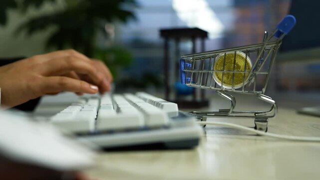Mini shopping cart with coin on desk, Miniature shopping cart with a gold coin near a computer keyboard and hands typing