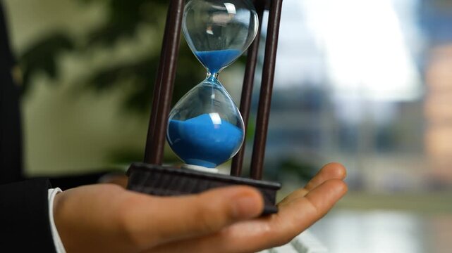 Hand holding an hourglass with blue sand in a wooden frame, blurred indoor background