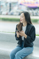 Fototapeta premium A beautiful Asian woman is walking along a city street with a smartphone and a backpack, posing with the city backdrop, and creating a social media post.