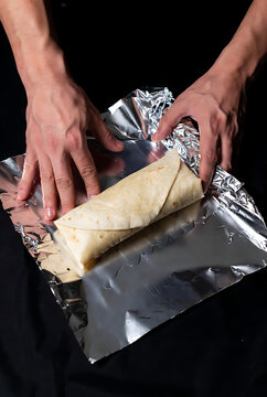 Hands wrapping fresh burrito with aluminum foil on dark background for food packaging or meal prep concept