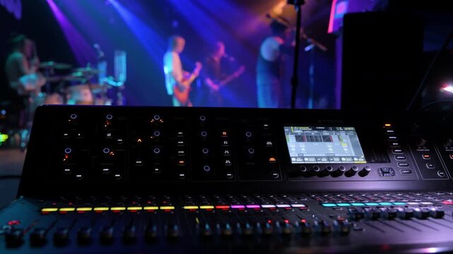 Professional audio mixing console in the foreground during a live music performance with band members performing on a colorful stage in the background. Sound engineer perspective at a concert venue.