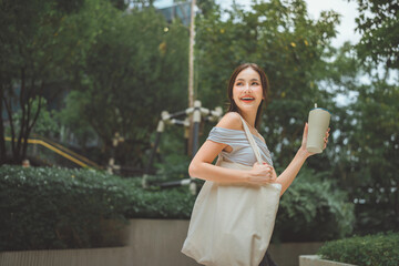 Happy Asian woman carrying reusable canvas tote bag and holding water bottle in the city, eco-friendly lifestyle and plastic-free concept, young stylish lady with fabric bag and refillable bottle