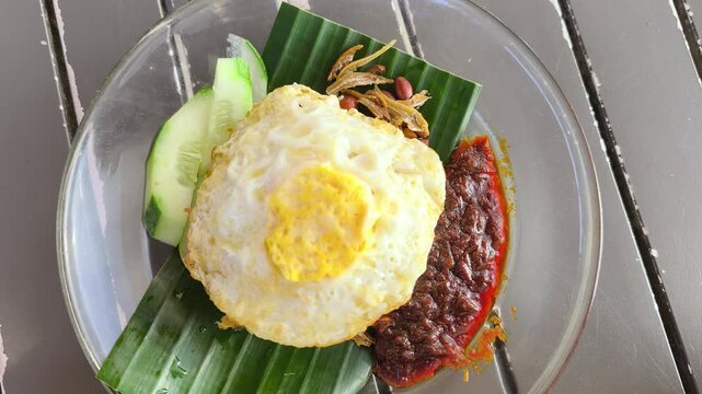 Fragrant nasi lemak is served on a glass plate with a fried egg. It comes complete with spicy sambal, crunchy peanuts, and fresh cucumber slices. This iconic Malaysian dish is ready to enjoy.