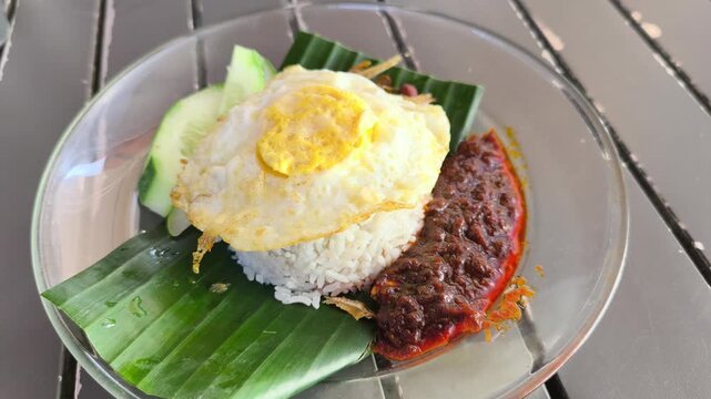 Fragrant nasi lemak is served on a glass plate with a fried egg. It comes complete with spicy sambal, crunchy peanuts, and fresh cucumber slices. This iconic Malaysian dish is ready to enjoy.