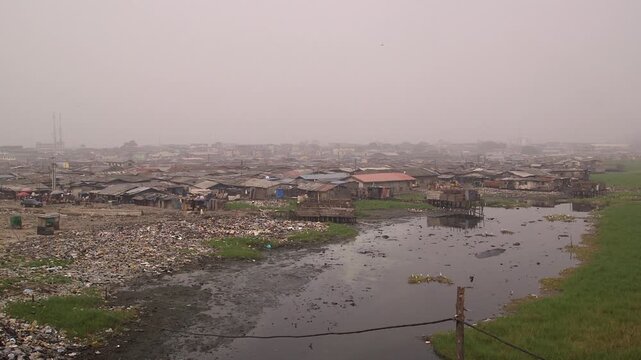 Hazy wide shot of a Lagos Lagoon waterfront settlement in Lagos, Nigeria, with shacks along polluted waterway and piles of trash, highlighting urban poverty and pollution.