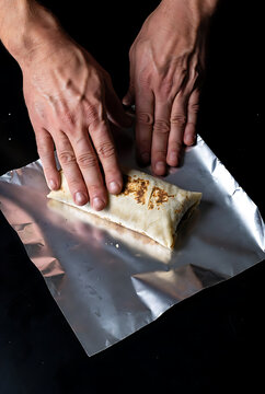 Hands rolling fresh burrito with seasoned meat and foil wrapper on dark kitchen counter for Mexican cuisine for Burrito Day celebration