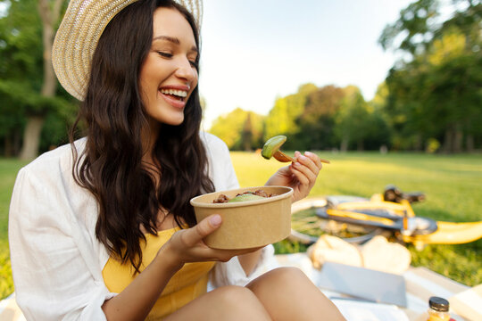 Fresh veggie meal. Happy woman eating from bowl and smiling, sitting on blanket in green summer park on sunny day, having snack during picnic
