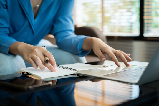 A woman is writing in a notebook and laptop. She is wearing a blue shirt and has a pen in her hand. The scene suggests that she is working on a project or writing something important