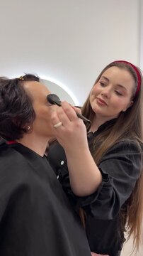 Professional makeup artist applying eye makeup cosmetics to female senior client during beauty treatment session in salon demonstrating MUA work technique for glamour makeover service preparation