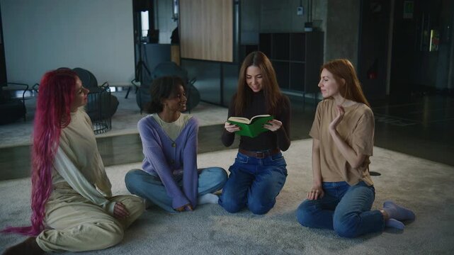 Four women sit on the floor in a room sharing thoughts during a group psychology session while one holds a book and speaks openly about feelings.