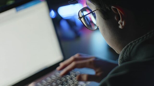 Over the Shoulder Shot of a Young Man with Glasses Typing on a Laptop at Night, Concept of Coding, Programming, or Cybersecurity