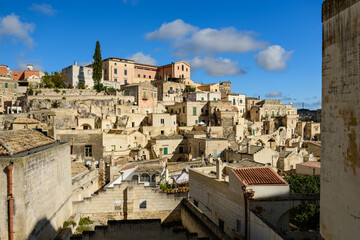 Naklejka premium Clustered stone houses and layered rooftops form a striking hillside scene in Matera, Italy, under bright sunlight and a vivid blue sky with scattered clouds.