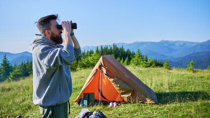 Man stands near orange tent, using binoculars to observe distant mountains. Grassy hilltop, under clear blue sky, lush green forests and rolling hills. Camping gear scattered around tent. © anatoliy_gleb