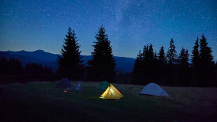Few tents on grassy clearing under dazzling, starry sky with Milky Way clearly visible. Tall pine trees and distant mountains frame scene, creating tranquil and awe-inspiring night in nature. © anatoliy_gleb