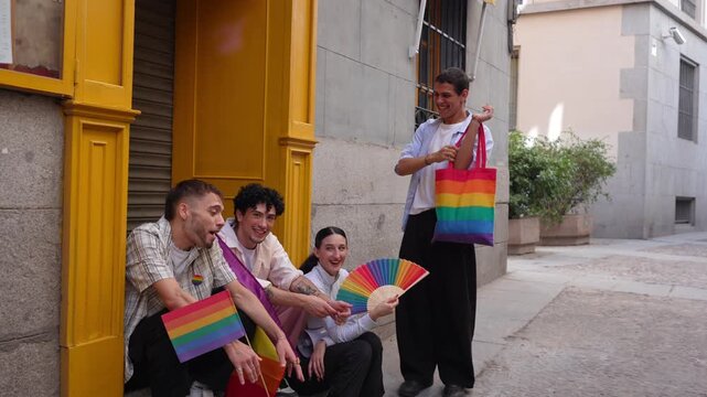 Happy group of friends chatting together on a city street. Diverse young people with rainbow flags, fan, and bag celebrating pride day