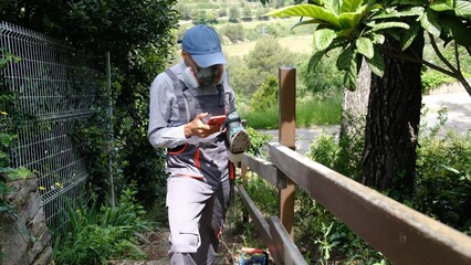 Man in protective respirator and work overalls using a smartphone, holding an electric sander, taking a break while renovating or repairing a wooden fence in a garden