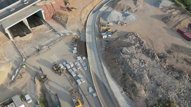 Aerial view of large highway construction site with heavy machinery, earthmoving, excavation and a new tunnel entrance amid dirt, gravel, concrete and staged building materials