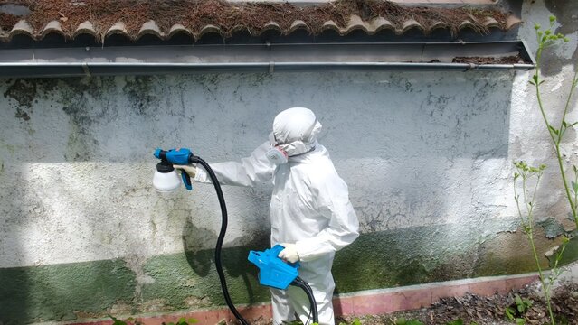 Person in protective suit spraying disinfectant on a dirty wall outdoors, performing sanitation against mold, viruses, or pests in a neglected building area