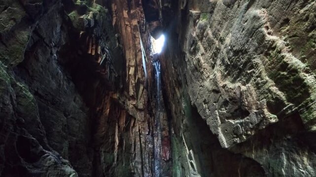A dramatic view from inside a narrow cave, showing a thin waterfall cascading down dark, mossy stone walls with bright daylight streaming through the opening above.