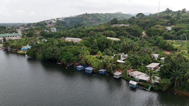 Cinematic drone shot of the Volta River bank in Akosombo, Ghana. Features traditional houses, floating platforms, and dense tropical vegetation under a cloudy sky. Nature and travel scene