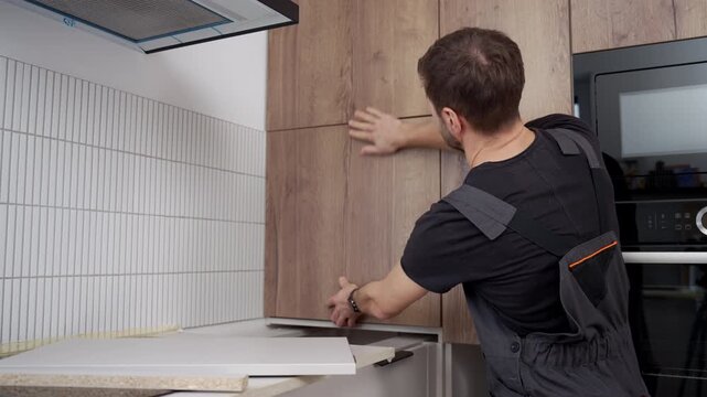 Man in overalls installing wooden kitchen cabinet door in modern apartment interior. Concept of home renovation, self made furniture assembly and repair service