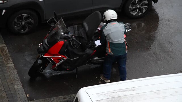 Moto y motorista consultando carpeta con papeles y documentos y abriendo maletero de asiento top case en d&iacute;a de lluvia