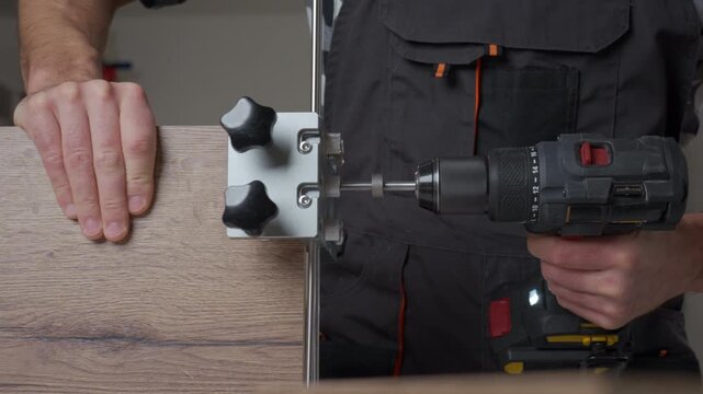 Close-up of carpenter drilling hole in wooden board using drill and metal jig tool. Man working in home workshop. Concept of woodworking and furniture assembly