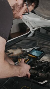 Vertical zoom shot of focused young male mechanic performing remote car engine inspection using endoscope with digital screen and camera, searching for malfunctions in gearbox, combustion or ignition